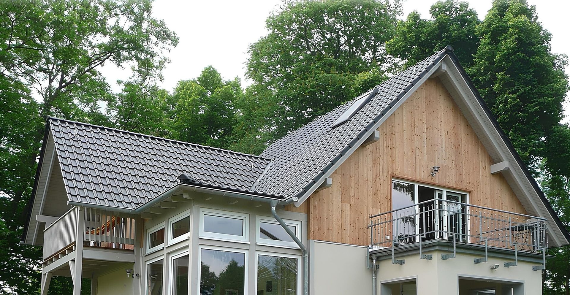 Modernes Einfamilienhaus mit Holz- und Putzfassade, dunklem Satteldach, großem Dachfenster und zwei Balkonen, umgeben von grünen Bäumen